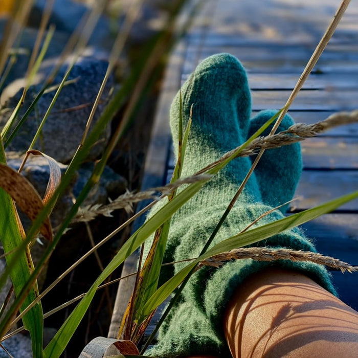 Teal knit socks worn with leg crossed over wooden dock surrounded by green marsh grasses.