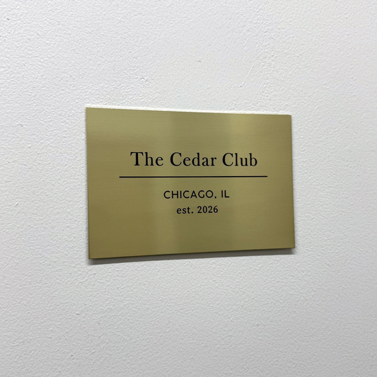 Brass gallery plaque with The Cedar Club lettering on a white wall.