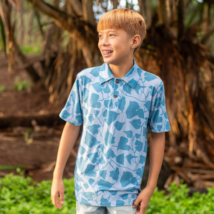 Child wearing a blue patterned shirt standing in a natural setting with trees and greenery.