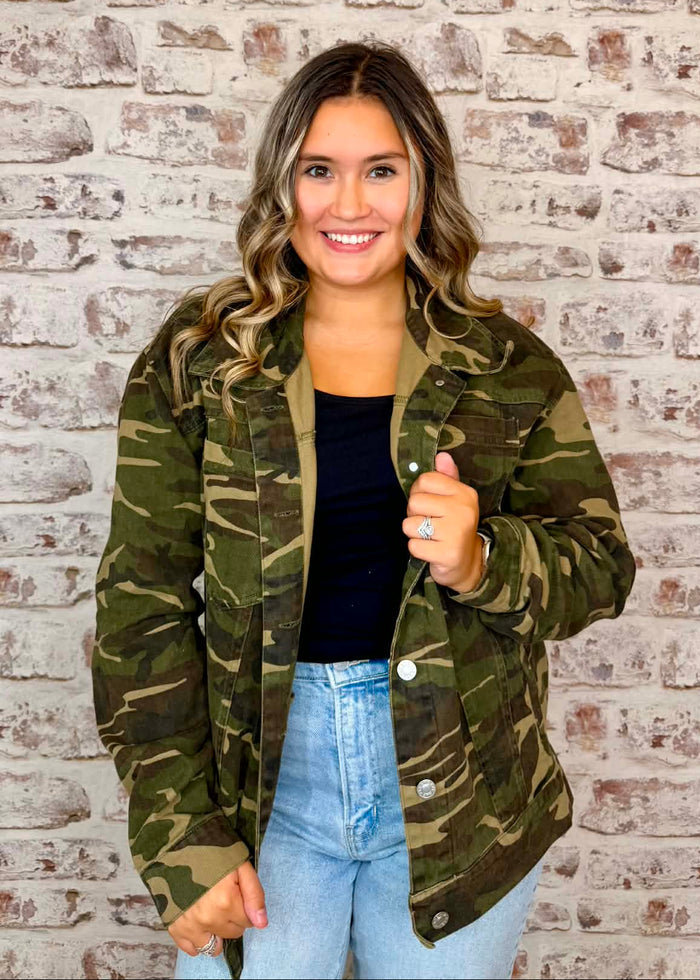 A woman wearing a green camouflage jacket over a black top and light blue jeans, standing in front of a white brick wall.