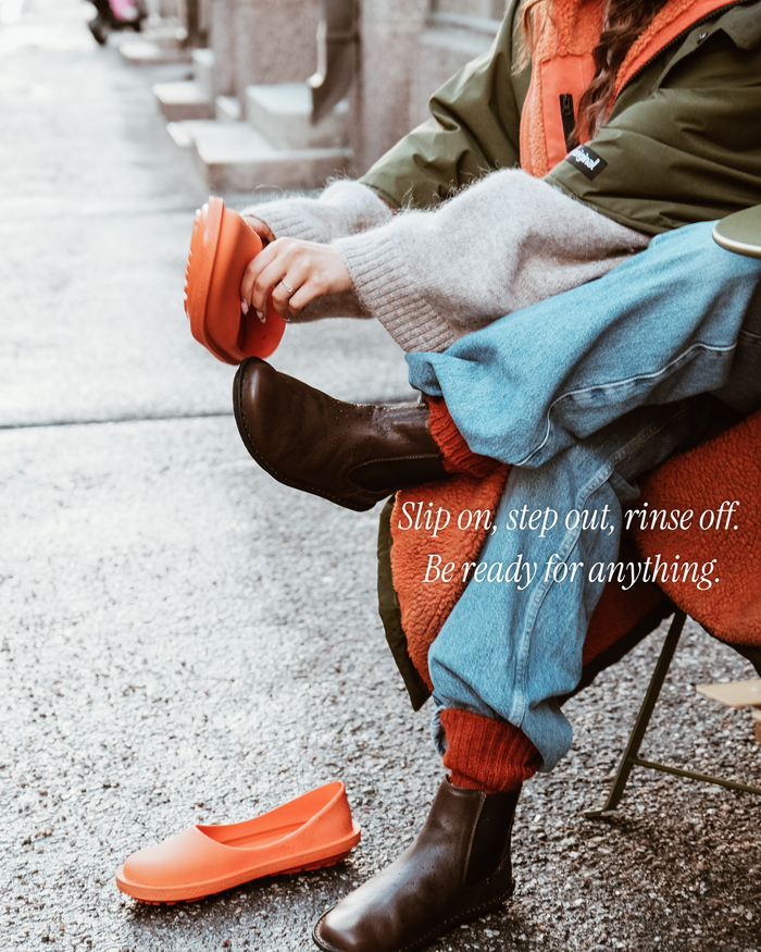 Person holding bright orange natural rubber slip-on shoe while seated outdoors.