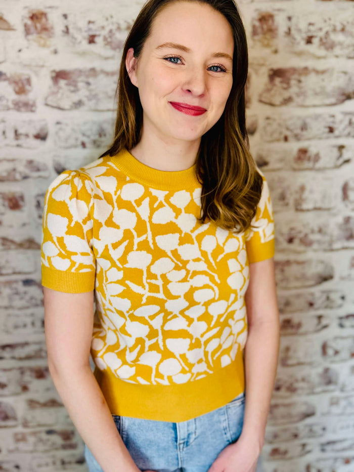 mustard yellow short sleeve sweater has a solid neckline and sleeve band color of mustard, and the print is imaginative of poppy flower silhouettes with a white in back neck button the color of a white pearl